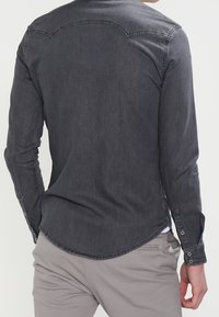 Man som bär en åtsittande mörkgrå långärmad tröja och ljusgrå byxor, sedd bakifrån mot en enkel bakgrund.