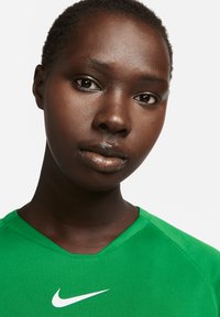 Grünes Nike Sportshirt mit V-Ausschnitt, ausgestattet mit einem weißen Swoosh-Logo. Glatte Textur, Nahaufnahme des Gesichts, welches den Hautton und die Merkmale hervorhebt.