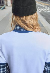 Bijela majica s plavim ovratnikom složena preko tamne karirane košulje, upotpunjena tamnim pletenim kapom, s teksturiranom kosom.