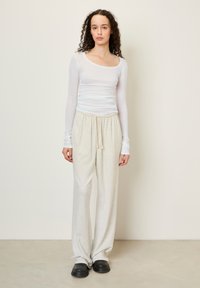 Top leggero bianco a maniche lunghe con scollo a barca, abbinato a pantaloni della tuta grigi chiari e larghi e ciabatte nere.