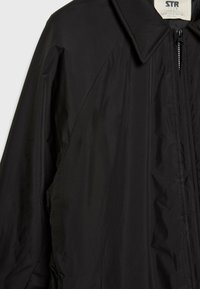 Giacca nera con chiusura a zip, grande colletto e texture liscia e leggera. Il design presenta spalle cadute e un leggero lucido.