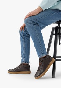 Sneaker alte marroni con tomaia testurizzata, punta tonda e suola in gomma a contrasto. Abbinati a jeans di un blu chiaro, seduto su uno sgabello.