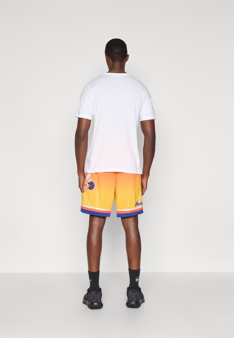 T-shirt bianca a maniche corte, abbinata a pantaloni da basket arancio-gialli con dettagli blu e un logo di lato. Sneakers nere.