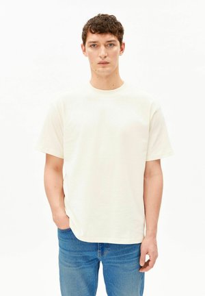 Jeune homme aux cheveux bruns bouclés et courts, portant un t-shirt blanc uni et un jean bleu, debout avec une main dans la poche.