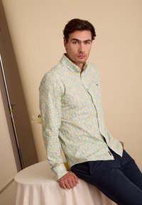 Lichtgele blouse met knopen en een bloemenpatroon in blauwe en groene tinten, gecombineerd met donkere broek. Model zit tegen een neutrale achtergrond.