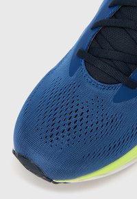Modrá športová obuv s textúrovaným sieťovinovým zvrškom, tmavými šnúrkami a neónovo zelenými doplnkami na podrážke. Pohľad zblízka na oblasť prstov.