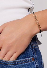 Złota bransoletka z łańcuchem na czarnym regulowanym sznurku, z utkwionymi ogniwami. Ustawiona na jasnej karnacji skóry z codziennymi dżinsami.