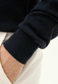 Marinblå tröja med ribbad mudd, mjukt material, figursydd design, med en synlig hand som vilar mot ljusfärgade byxor.