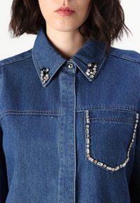 Denimskjorta med klassisk krage, försedda med ädelstensdetaljer på kragen och en dekorativ ficka. Blått tyg, knapprad fram.