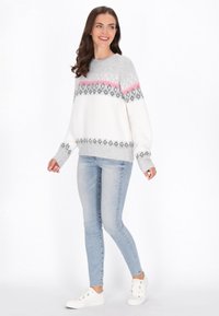 Suéter blanco con patrones geométricos en gris y rosa, de mangas largas, cuello redondo, combinado con jeans azul claro y zapatillas blancas.