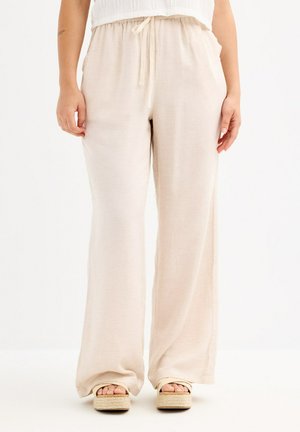 Persona vistiendo pantalones sueltos de color beige claro con cordón y sandalias beige con plataforma, de pie frente a un fondo blanco liso.