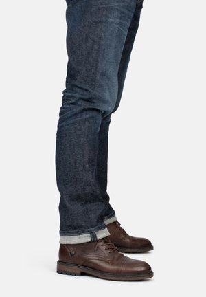 Stivali marroni in pelle con lacci e finitura texturizzata, abbinati a jeans denim scuri, con risvolti arrotolati e vestibilità slim.