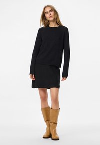 Nő fekete hosszú ujjú pulóver és szoknya szettben, barna térdig érő bőr csizmával, sima fehér háttér előtt állva.