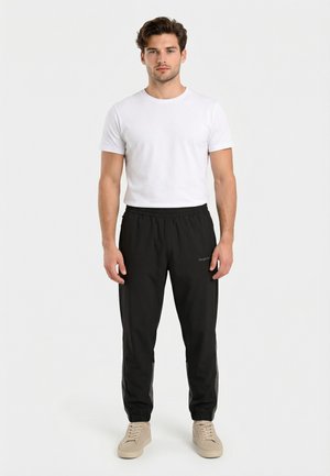 Uomo in piedi di fronte, che indossa una t-shirt bianca a maniche corte, pantaloni jogger neri e sneakers beige su uno sfondo neutro.
