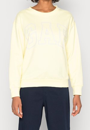 Personne portant un sweat-shirt jaune pâle avec un lettrage discret "GAP" et un pantalon bleu foncé, debout devant un fond clair uni.