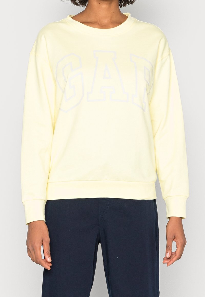 Personne portant un sweat-shirt jaune pâle avec un lettrage discret "GAP" et un pantalon bleu foncé, debout devant un fond clair uni.