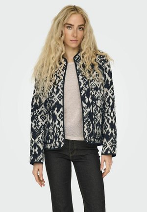 Veste à motif bleu marine et blanc en tissu avec une finition texturée. Elle possède un col arrondi et une fermeture éclair, portée sur un haut léger.