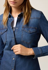 Camicia di jeans di colore blu scuro con colletto classico, due tasche sul petto e chiusura con bottoni. Presenta una texture leggermente sbiadita.