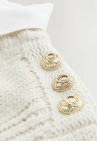 Kötött pulóver törtfehér színben, oldalán három kör alakú, fonott arany gombbal, bordázott textúrával és sima anyagból készült gallérral.