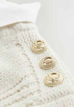 Sweter dzianinowy w kolorze ecru z trzema okrągłymi, plecionymi złotymi guzikami po bokach, o prążkowanej fakturze i kołnierzu wykonanym z gładkiego materiału.