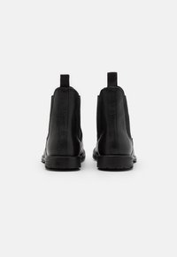 Botas de tobillo de cuero negro vistas desde atrás, con tiradores y suelas texturizadas sobre un fondo blanco.