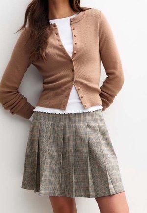 Frau trägt eine geknöpfte Kamelhautfarbene Strickjacke über einem weißen Oberteil, kombiniert mit einem hoch taillierten, plissierten karierten Rock vor weißem Hintergrund.
