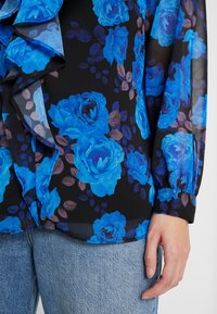 Blouse noire transparente à motif de roses bleues et bordure à volants, portée avec un jean en denim bleu clair, main reposant sur le côté.