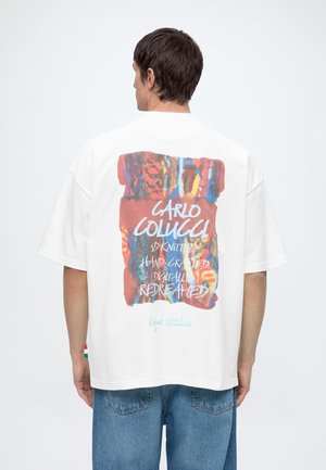 Mann trägt weißes, übergroßes T-Shirt mit buntem abstraktem Motiv und Text auf dem Rücken, kombiniert mit blauer Jeans vor einfachem Hintergrund.