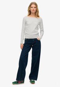 Grauer, langärmeliger, schulterfreier Oberteil kombiniert mit dunkelblauen, weiten Jeans und grünen Turnschuhen mit rosa Akzenten.