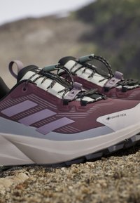 Lila-weiße Gore-Tex Trailrunning-Schuhe mit schwarzen Schnürsenkeln auf felsigem Außenboden, die profilierte Sohlen und Markestreifen zeigen.
