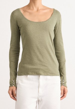 Femme portant une chemise à manches longues vert olive avec un col rond et des détails de boutons près des poignets, associée à un pantalon blanc.