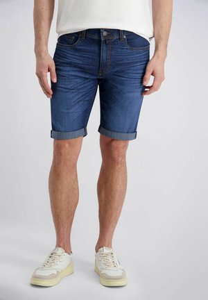 Mann trägt hochgekrempelte blaue Jeansshorts, weiße Sneakers und ein weißes Hemd vor einem schlichten hellen Hintergrund.