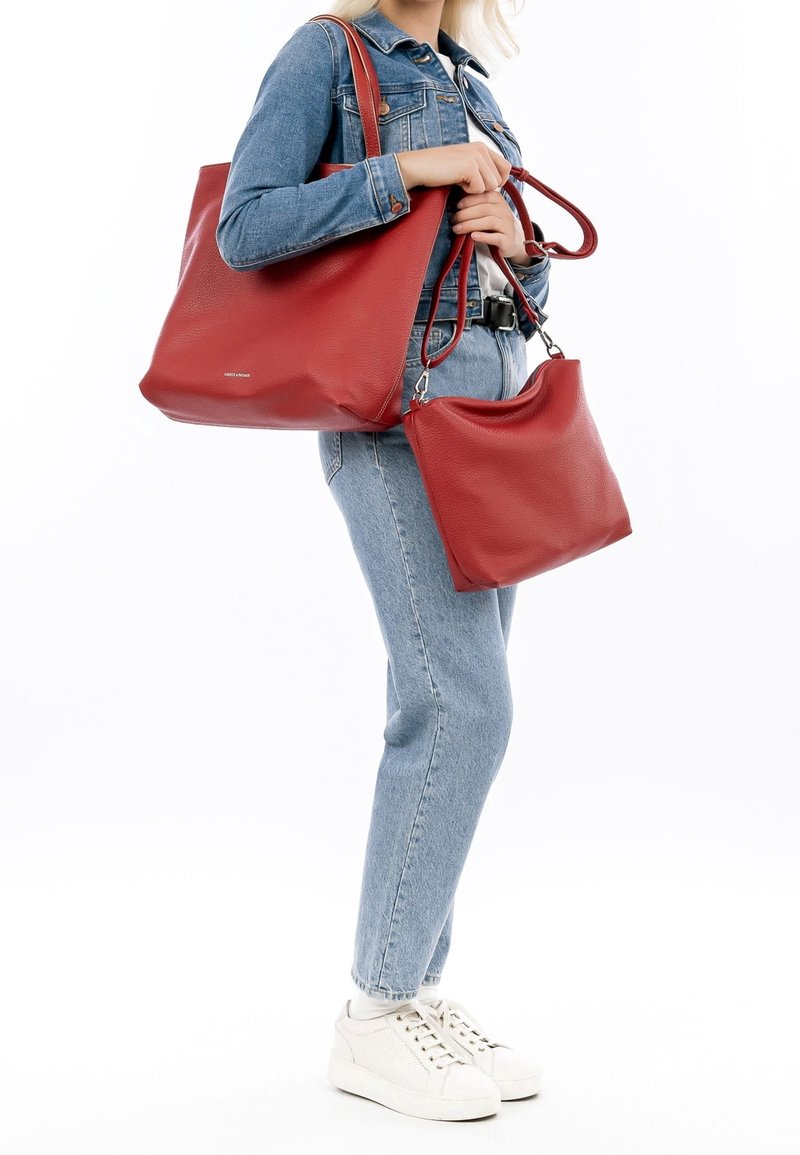 Femme en tenue en jean bleu tenant deux sacs à main en cuir rouge, un porté à l'épaule et un tenu en main, chaussée de baskets blanches.