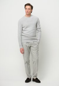 Helles graues Sweatshirt mit strukturiertem Strick, kombiniert mit hellgrauen, maßgeschneiderten Hosen. Modell trägt dunkle Slipper und steht vor einem schlichten Hintergrund.