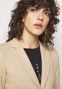 Blazer beige avec revers, porté sur une chemise noire ornée d'accents dorés. Cheveux bouclés et boucles d'oreilles dorées pendantes complètent le look.