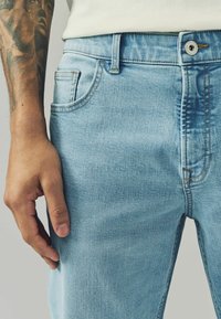 Jeans din denim de culoare albastră deschisă cu un design ajustat, prezentând buzunare frontale, cusături vizibile și o închidere cu un singur nasture. Textură moale.