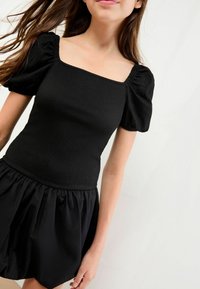 Schwarzes Kleid mit einem gerafften Oberteil, quadratischem Ausschnitt und puffärmeligen kurzen Ärmeln, kombiniert mit einem fließenden, gerafften Rock.