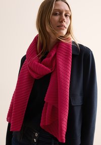 Lyserød ribbestrik scarf snoet omkring halsen, lavet af et blødt materiale, der hænger med et tekstureret mønster mod en mørk jakke.