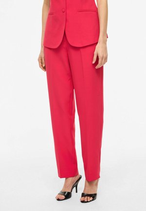 Femme portant un pantalon rouge ajusté et un blazer assorti avec des sandales à talons hauts noires ouvertes à l'avant, debout devant un fond blanc.