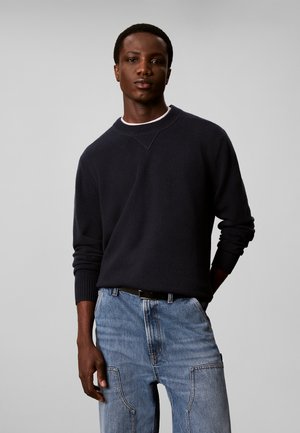 Ung mand iført en mørk langærmet sweater og lyseblå jeans står med den ene hånd delvist i lommen mod en ensfarvet baggrund.