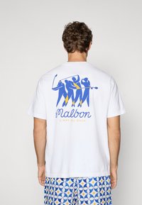 Hvid bomulds t-shirt med et blåt og gult grafisk design af fire figurer, der spiller golf, med teksten "Malbon" og "L'Art Du Coup" nedenfor.