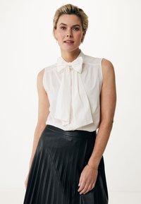 Blusa bianca senza maniche con un grande fiocco al colletto; abbinata a una gonna nera plissettata con una cintura liscia.