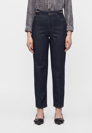 Personne portant un jean bleu foncé taille haute, coupe droite, avec des détails de boucles sur les côtés, un t-shirt gris à manches longues et des chaussures noires pointues.