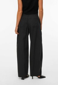 Žena oblečená v volných černých kalhotách, černém tílku a černých podpatcích, stojící zády k divákovi na jednobarevném bílém pozadí.