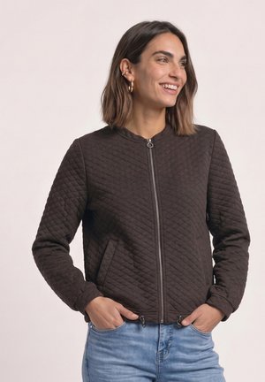 Frau mit kurzen braunen Haaren, lächelnd, trägt goldene Creolen, eine dunkelbraune gesteppte Reißverschlussjacke und hellblaue Jeans, Hände in den Taschen.