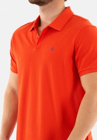 Camiseta polo naranja hecha de tela texturizada con cuello clásico, que presenta un pequeño logo azul en el lado izquierdo del pecho y mangas cortas.