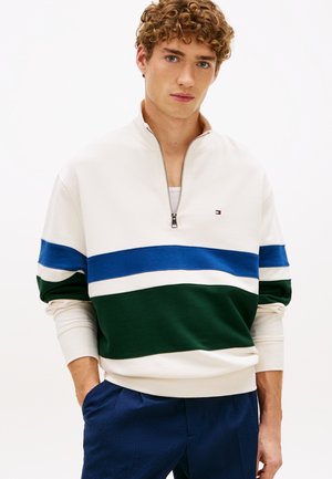 Młody mężczyzna z kręconymi włosami w białym swetrze z niebiesko-zielonymi poziomymi paskami i granatowych spodniach na jednolitym tle.