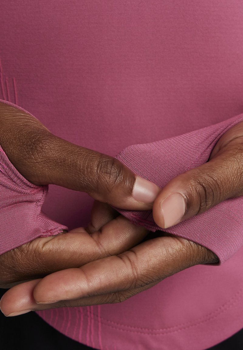 Manos de piel oscura ajustando el agujero del pulgar de una camisa de manga larga rosa contra un fondo de tela rosa.