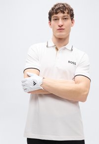 Λευκό πόλο μπλουζάκι με μαύρες και χρυσές λεπτομέρειες, που φέρει λογότυπο και κοντά μανίκια. Το μοντέλο φοράει λευκό γάντι γκολφ με μαύρες λεπτομέρειες.