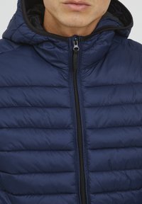 Námornícka modrá prešívaná bunda s kapucňou, s horizontálne prešívanými časťami, predným zipsom a čiernym podšitím okolo kapucne.